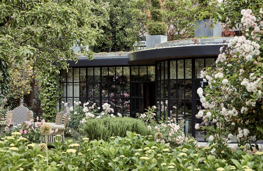 A view of the garden pavilion from the middle of the garden. White iceberg roses and flowering jasmine are visible at...