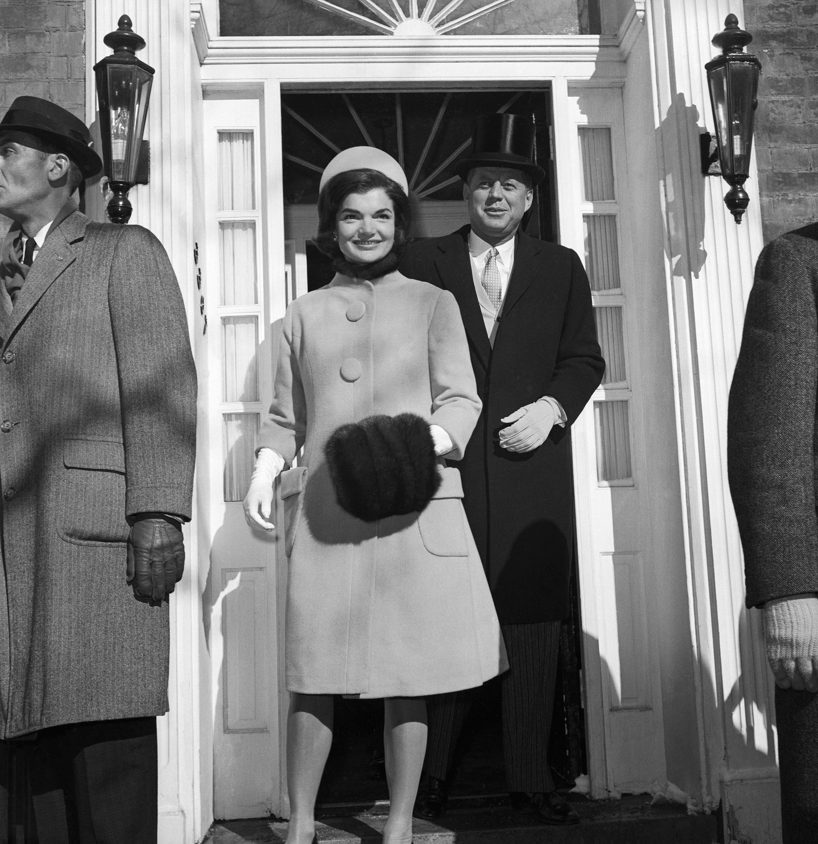 Jackie Kennedy in jacket and hat smiling standing to left JFK in tap hat and long jacket standing to right behind here...