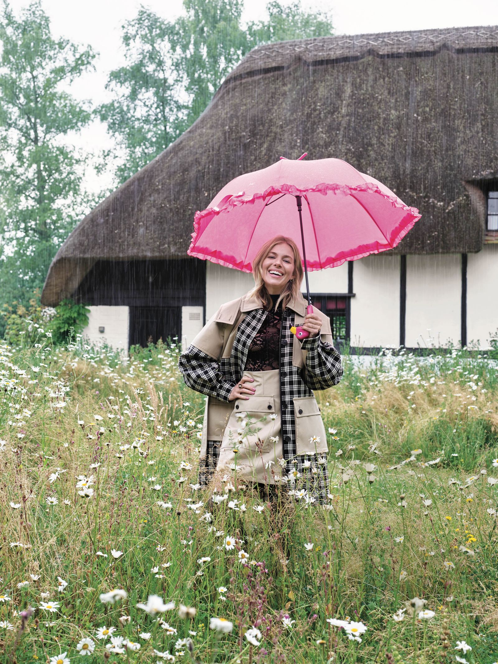 Sienna Miller with pink umbrella standing in meadow of her home in the rain home in background