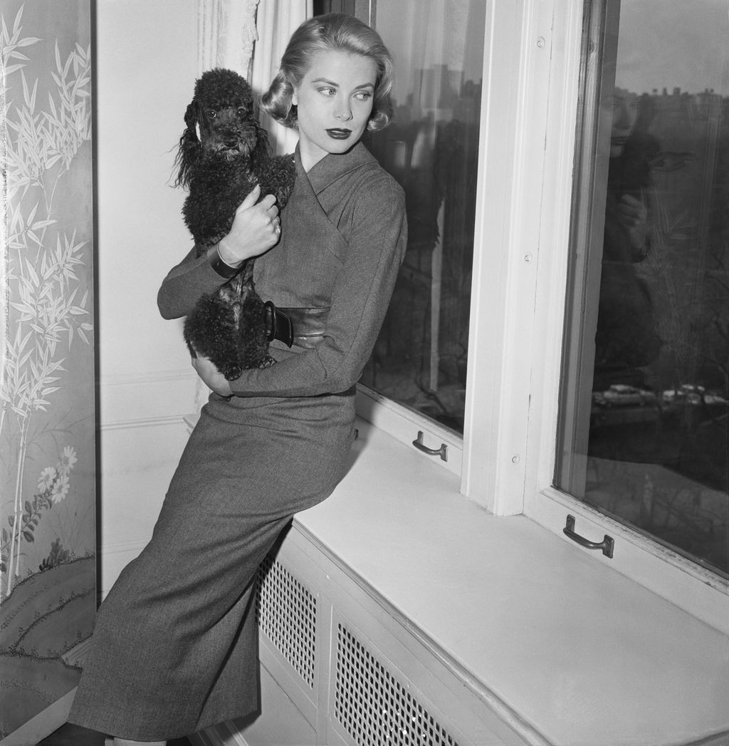blackandwhite photo of Grace Kelly at home holding black dog sat on bench beneath window to right leafy wallpaper to left