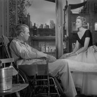 black-and-white photo in Rear Window set of Jimmy Stewart, left, sitting in wheelchair with left leg extended in cast; Grace Kelly seated facing him on window ledge; windows open to reveal city set of apartments across courtyard, skyline; to left, bucket with a bottle inside atop a chair